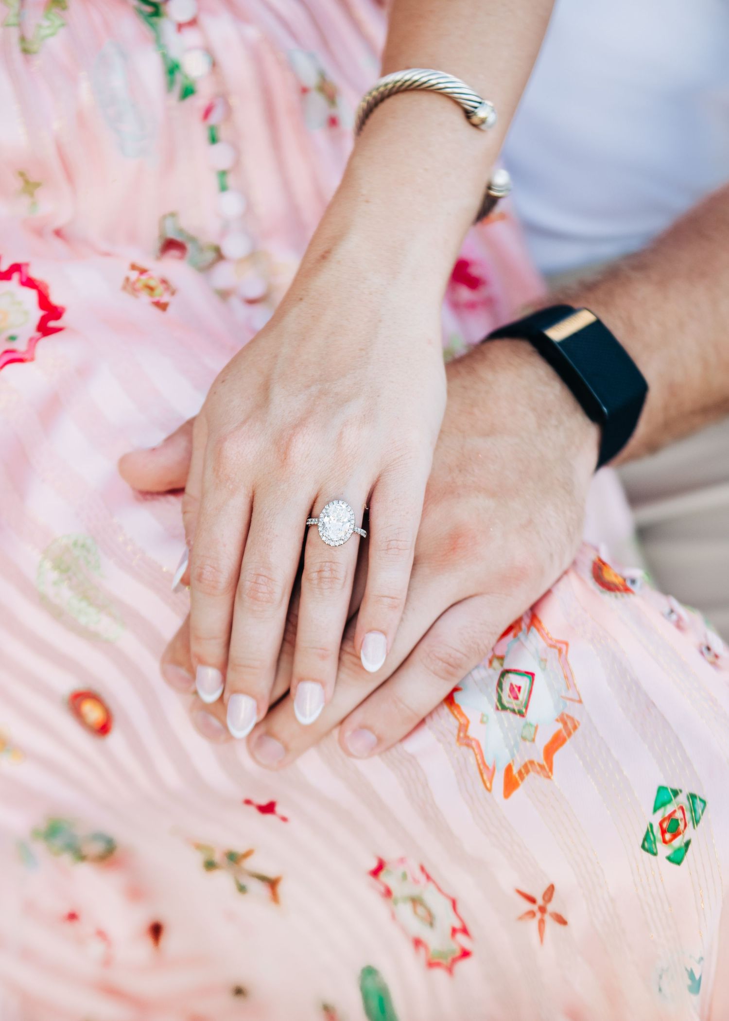 Close-up of couple's hands showing diamond engagement ring, silver bracelet, and black fitness tracker on pink floral dress.