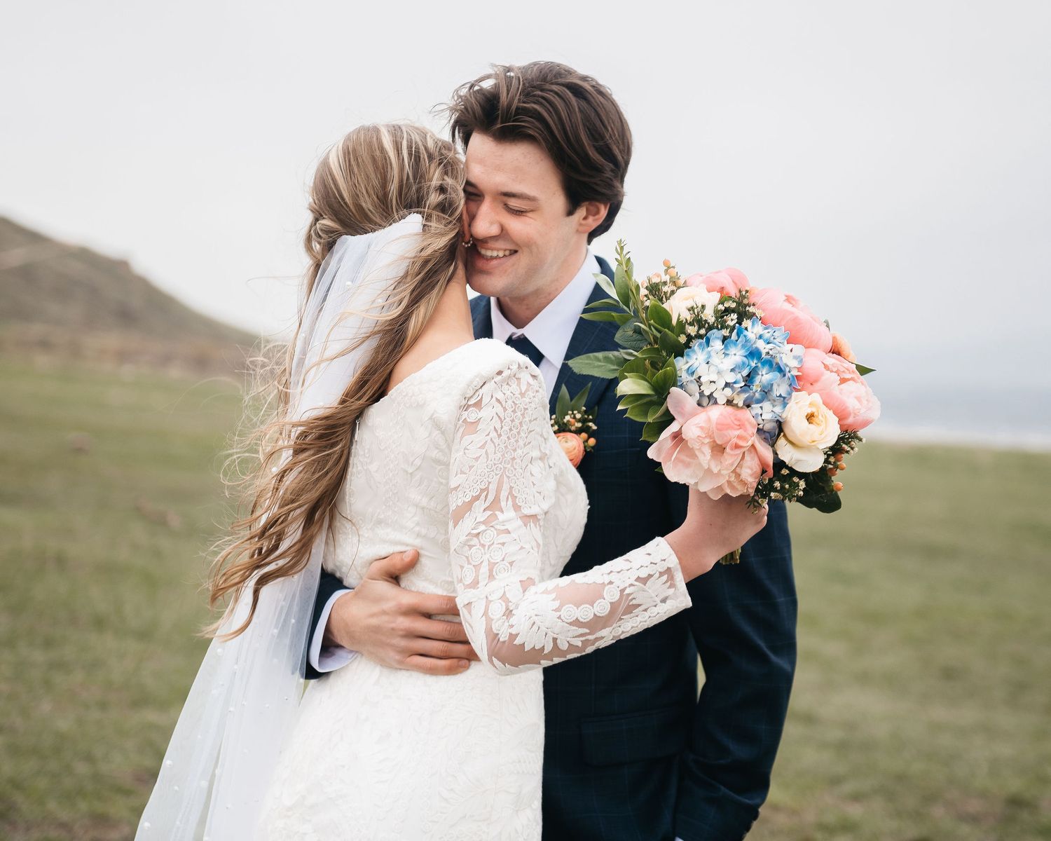 A smiling groom in a navy suit embraces his bride in a white lace dress, holding a pink and blue bouquet outdoors.