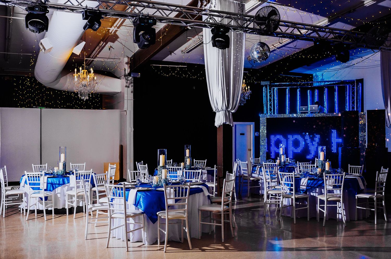 An elegant reception venue with blue lighting, white chairs, and chandeliers set up for a formal event.