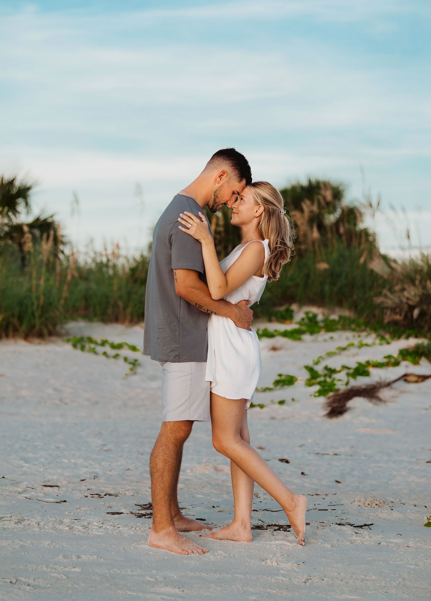 Couple embracing and smiling on a sandy beach with palm trees, wearing casual summer outfits.