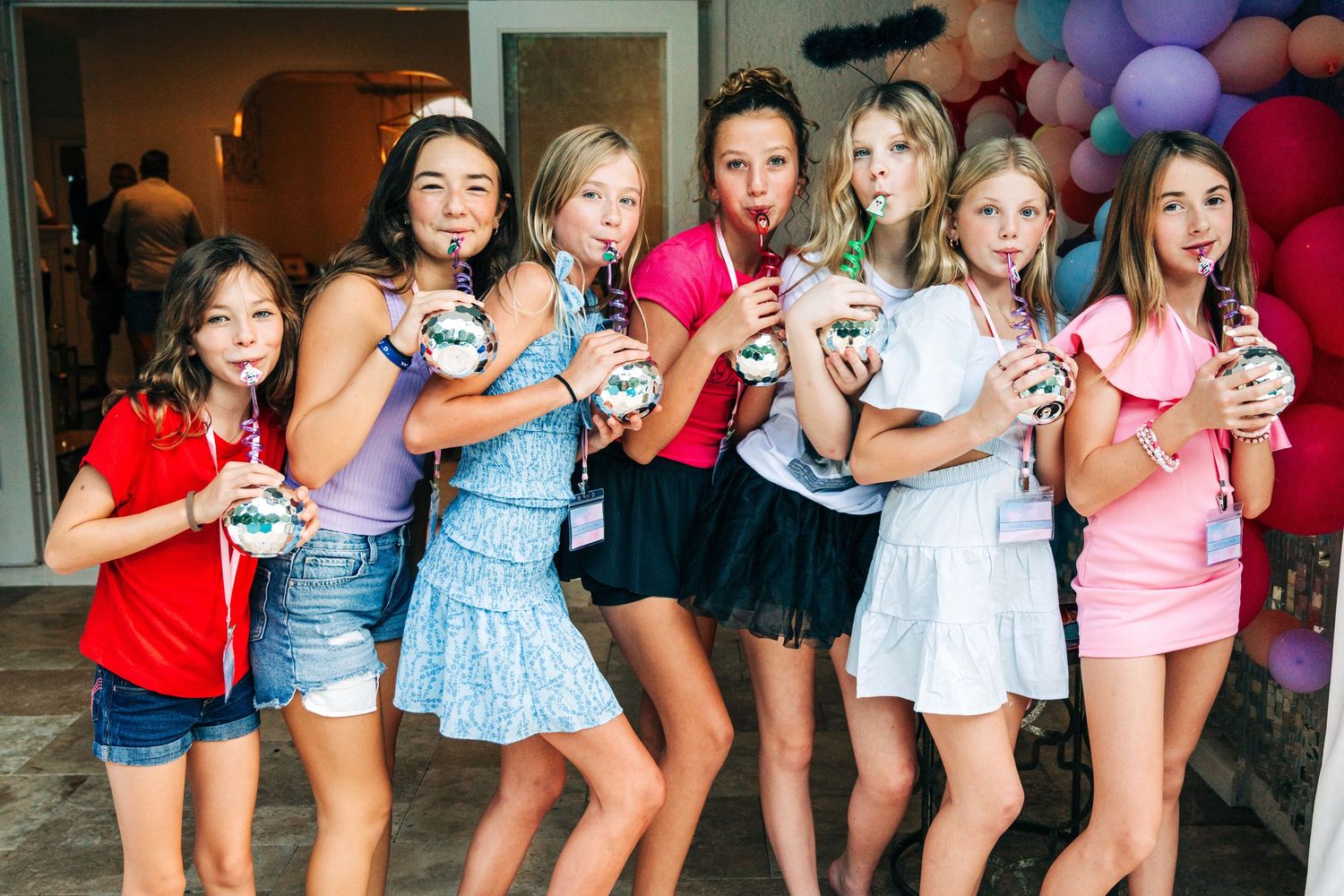 Group of young girls posing together at a colorful birthday party, holding drinks and smiling outdoors.