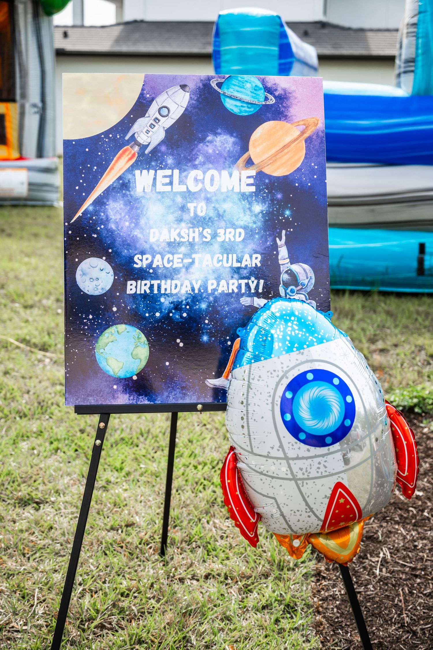 Space-themed 3rd birthday party welcome sign on easel with rocket balloon decoration outdoors.
