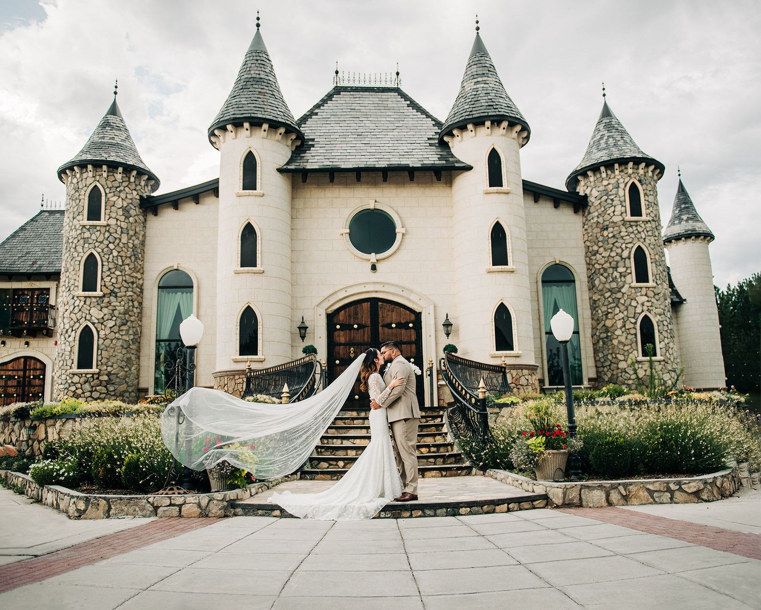 Bride and groom kissing in front of a fairy-tale castle wedding venue with stone towers and flowing veil.