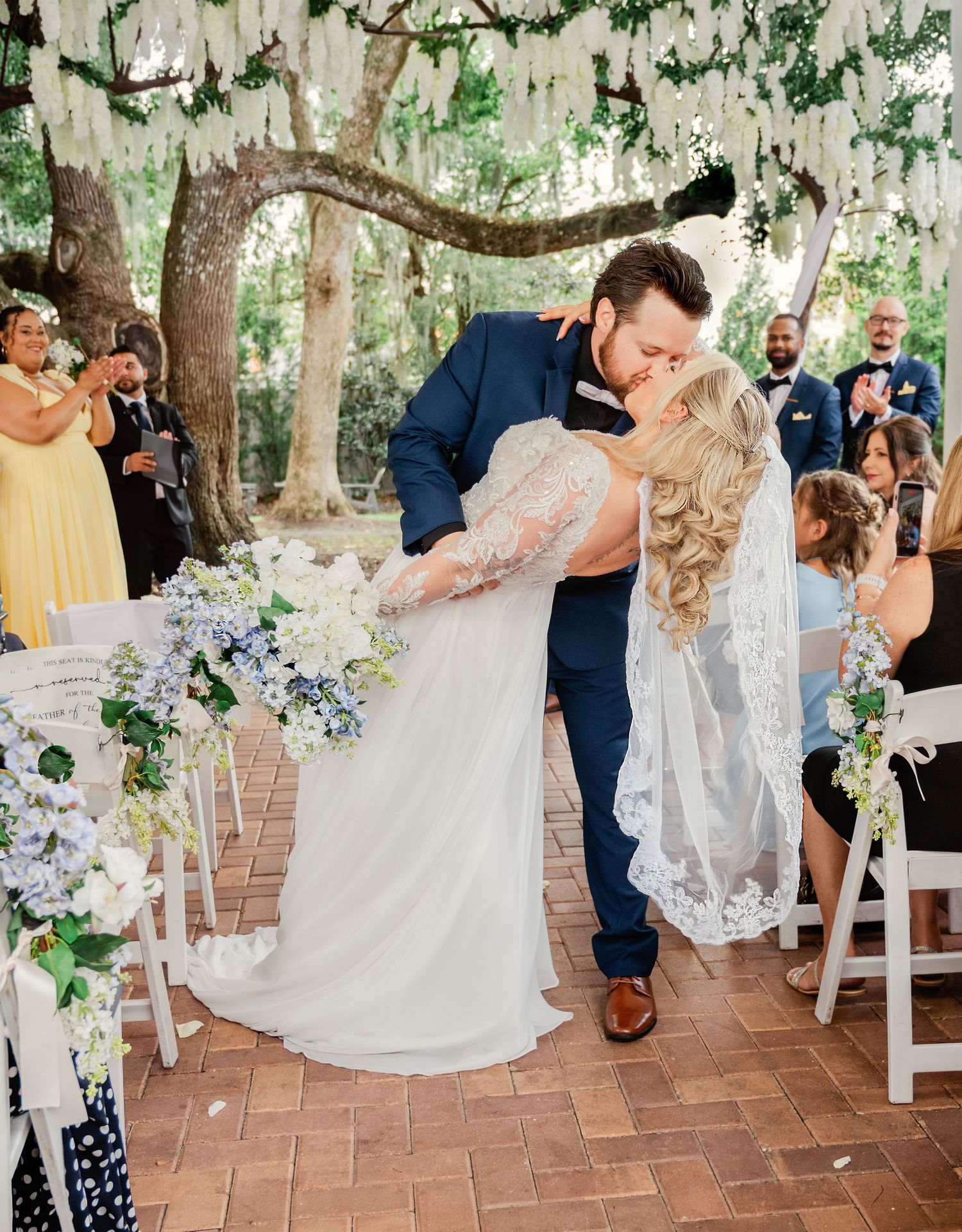 Bride and groom share first kiss at outdoor wedding ceremony under flowering tree with blue and white floral arrangements.