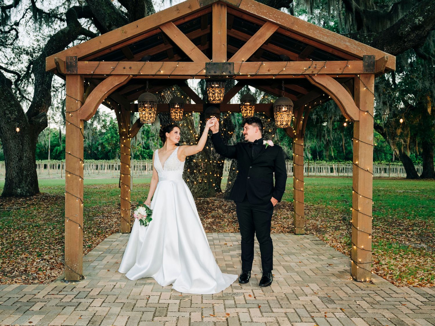 Bride and groom dancing under a rustic wooden gazebo with hanging lanterns at an outdoor wedding venue.