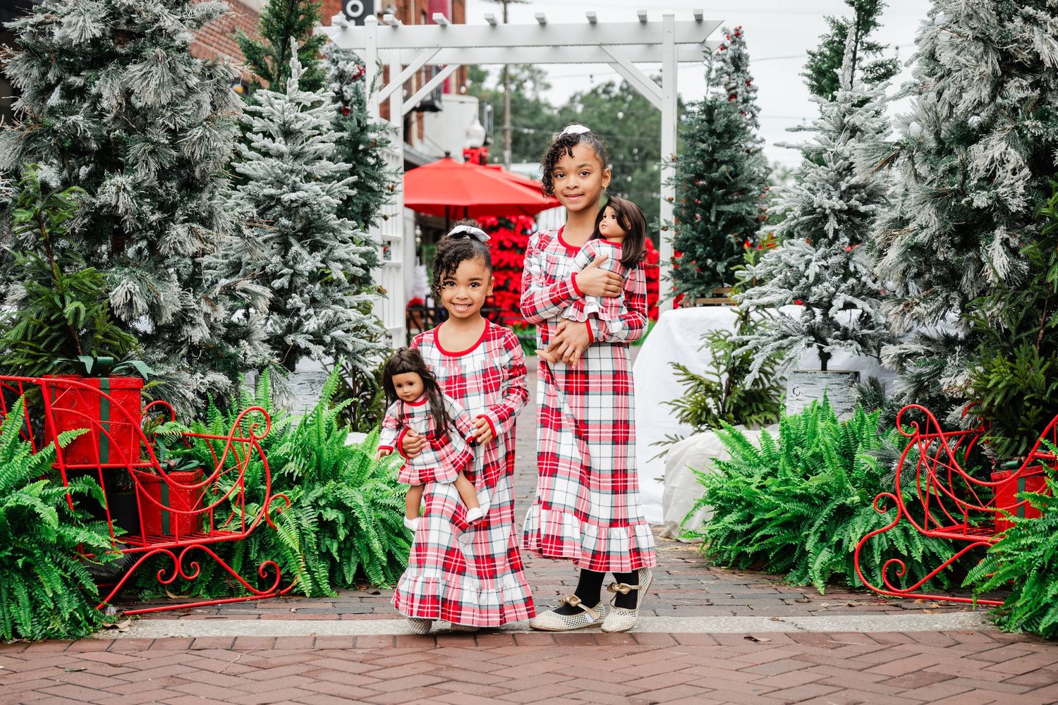 Two girls in matching red plaid Christmas pajamas hold dolls in festive holiday outdoor decor with evergreen trees.