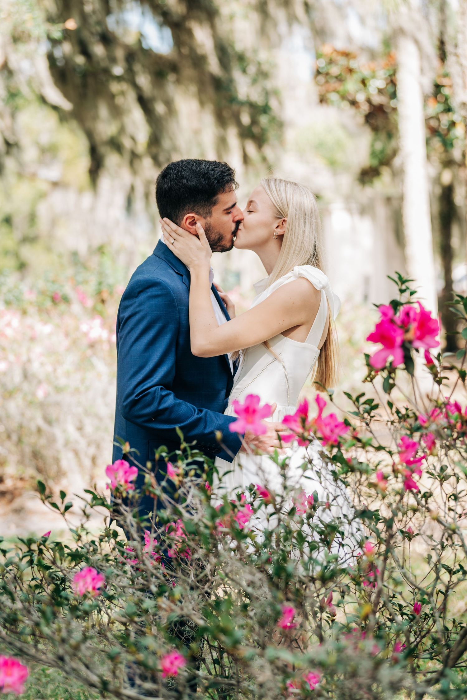Couple sharing a romantic kiss surrounded by vibrant pink azalea blooms in a lush garden setting.