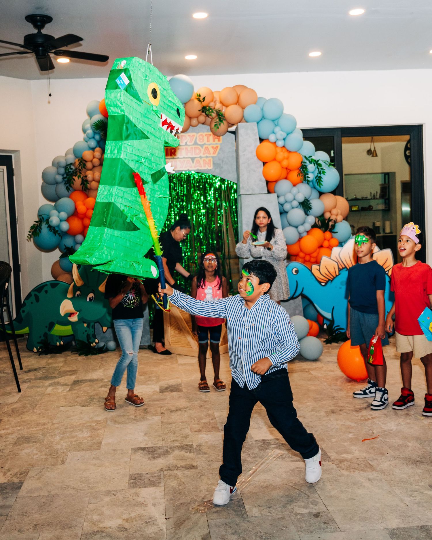 Boy swinging at a dinosaur pinata at a colorful dinosaur-themed birthday party with balloon arch decor.