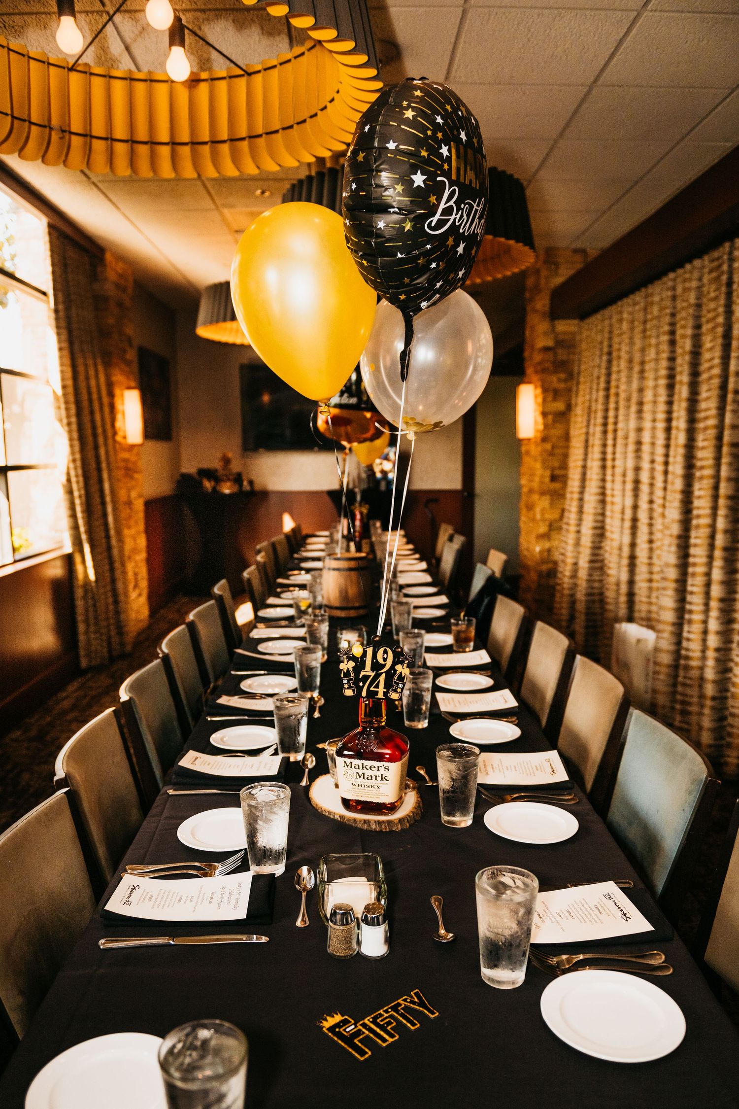 Elegant birthday party table setup with black tablecloth, gold and black balloons, white plates, and Makers Mark whiskey centerpiece.