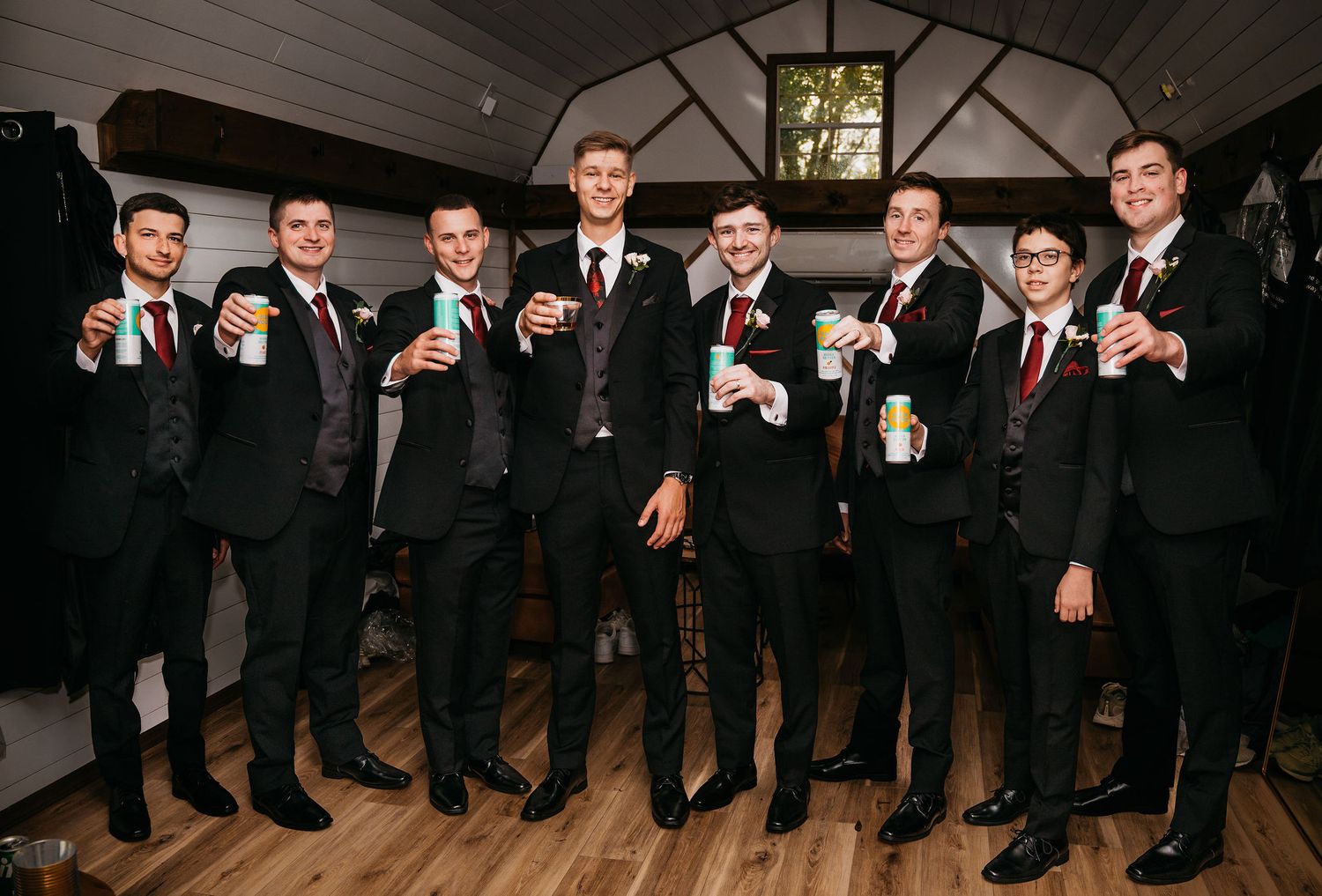 Eight groomsmen in black suits with boutonnieres raising drinks in a toast inside a rustic wedding venue.