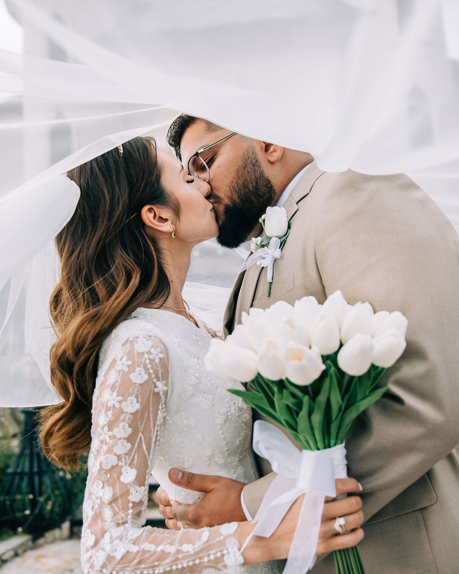 Bride and groom share a romantic kiss under a flowing veil, bride holding white tulip bouquet in lace wedding dress.