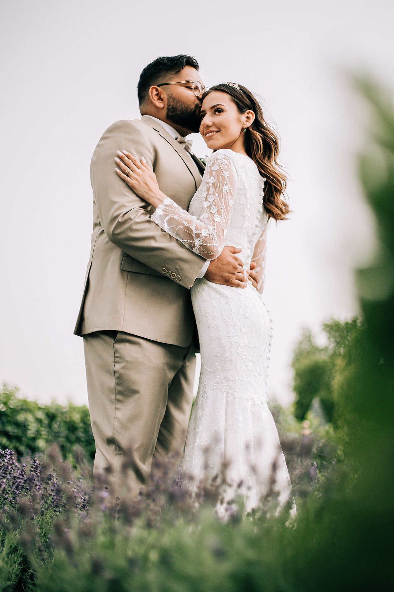 Bride in lace gown and groom in tan suit embrace in lavender field, sharing a tender kiss outdoors.