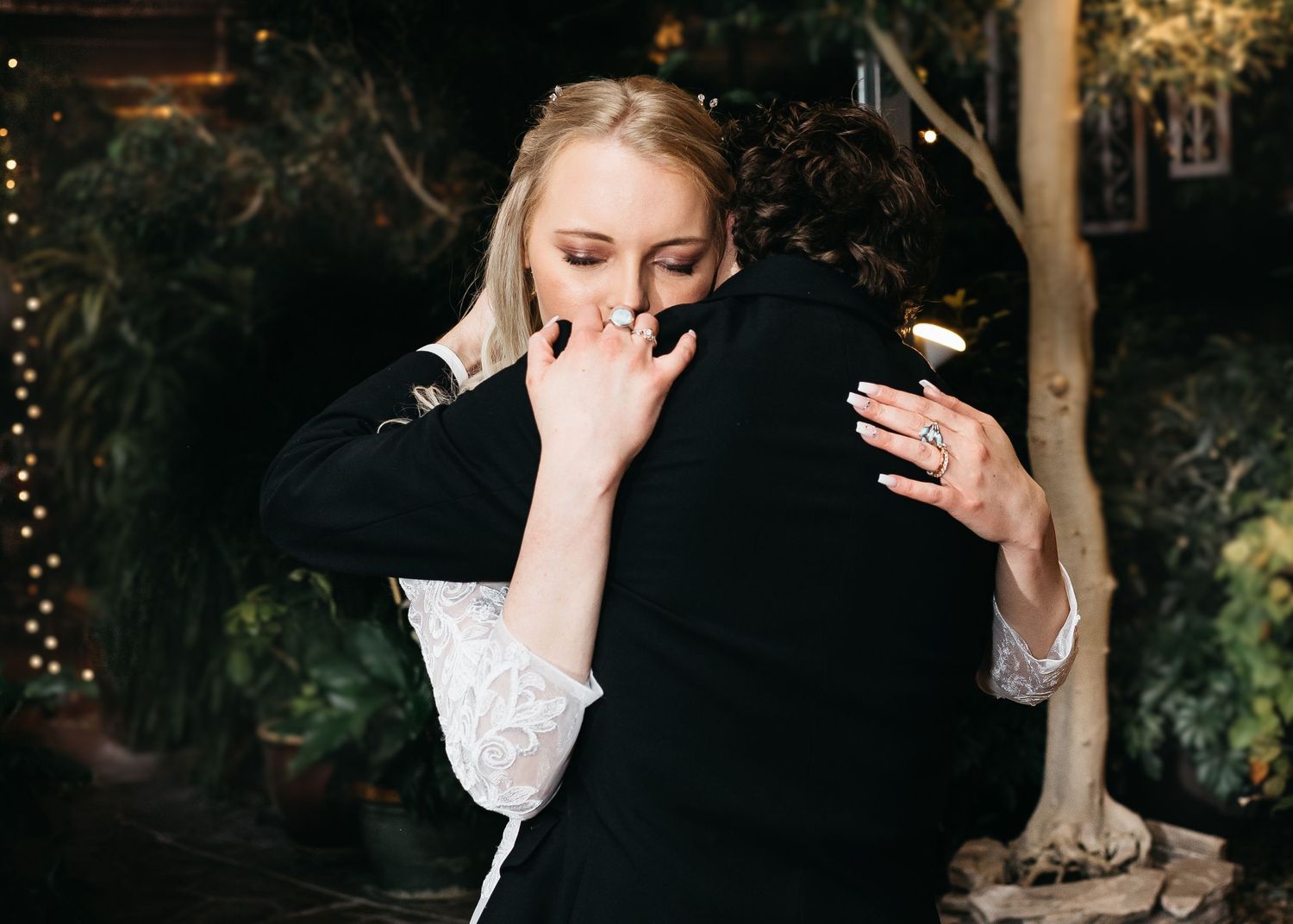 Emotional couple sharing a heartfelt hug at an indoor garden wedding reception with warm fairy lights.