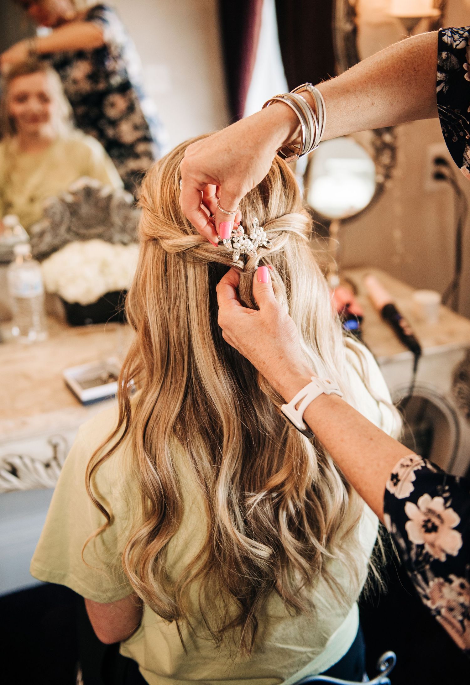 Stylist adding floral accessories to a half-up braided hairstyle on a blonde bride during wedding preparations.