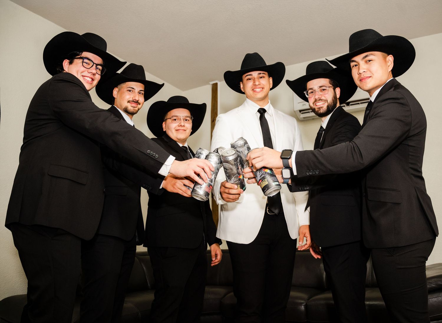 Six men in black suits and cowboy hats toasting with silver cups, one in white suit, at a formal celebration.
