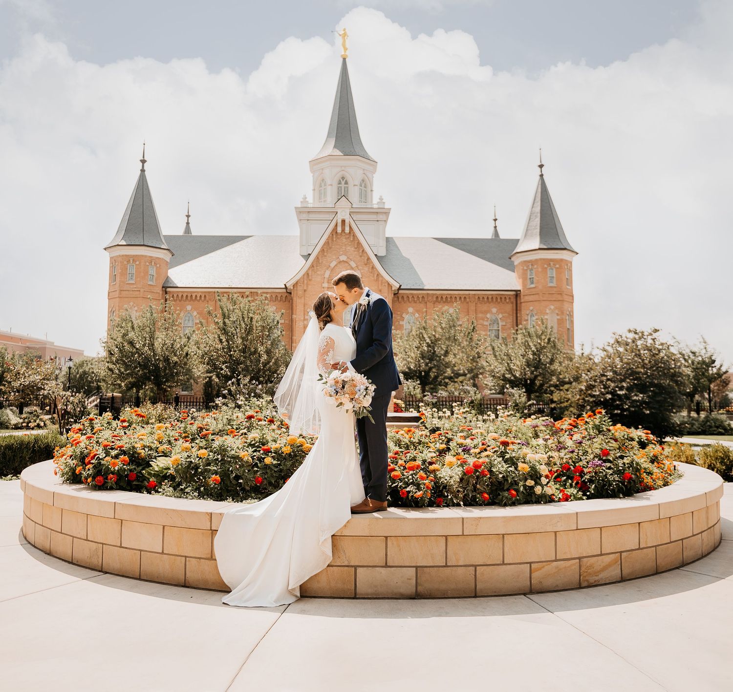 Bride and groom pose in front of a brick LDS temple surrounded by colorful flower gardens on their wedding day.