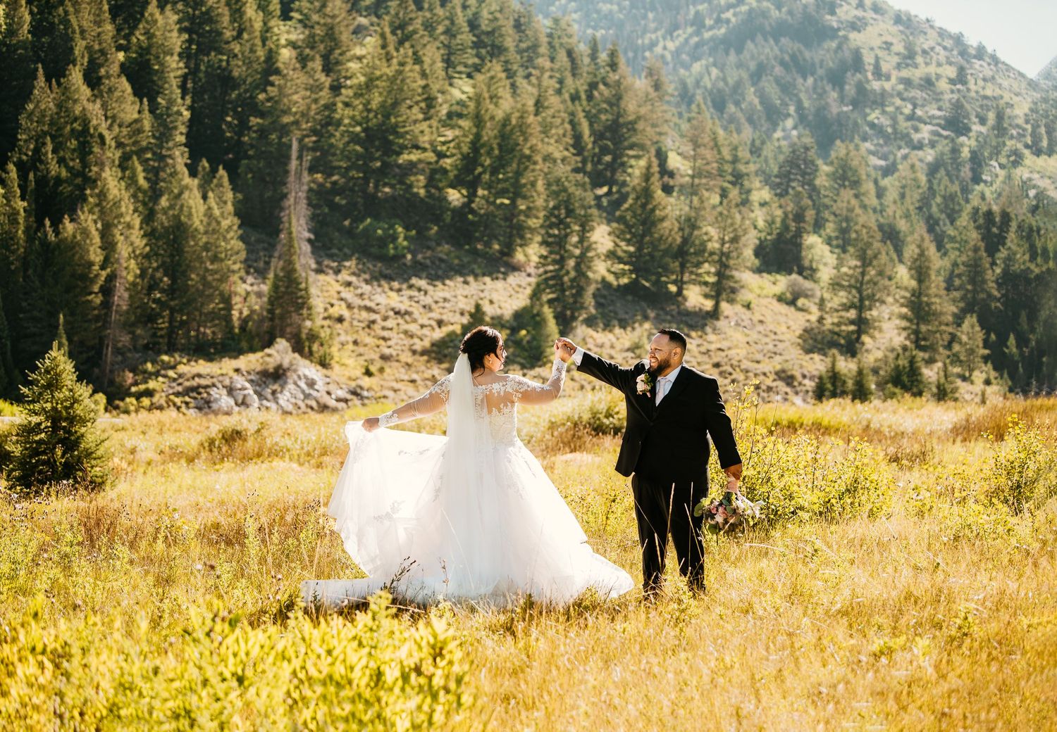 Bride and groom dancing in golden meadow with pine forest and mountain backdrop on their wedding day.