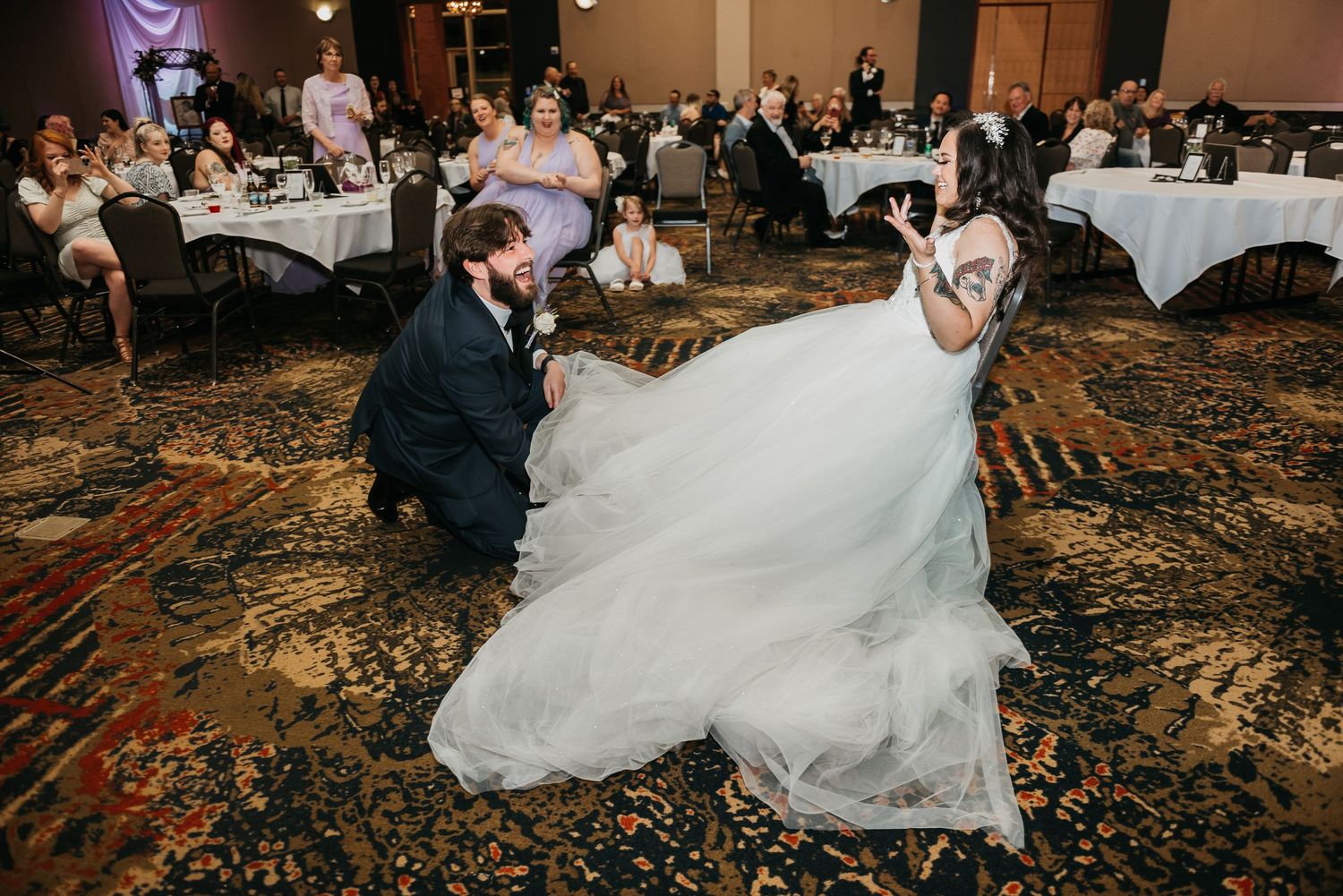 Groom kneeling before bride in white gown during wedding reception garter removal on patterned ballroom floor.