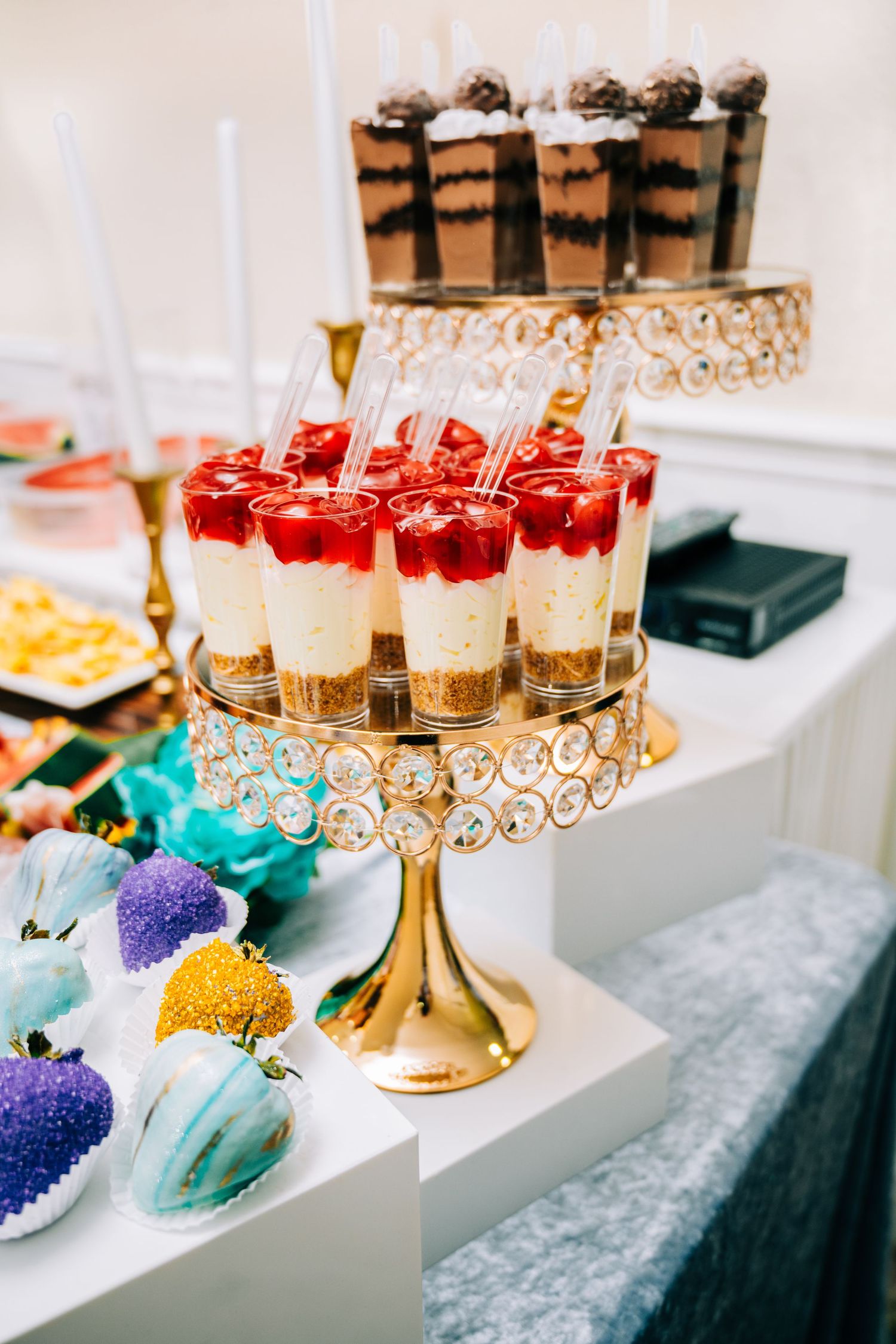 Elegant dessert table with layered cheesecake cups on gold stand, chocolate parfaits, and colorful cake pops at a party.