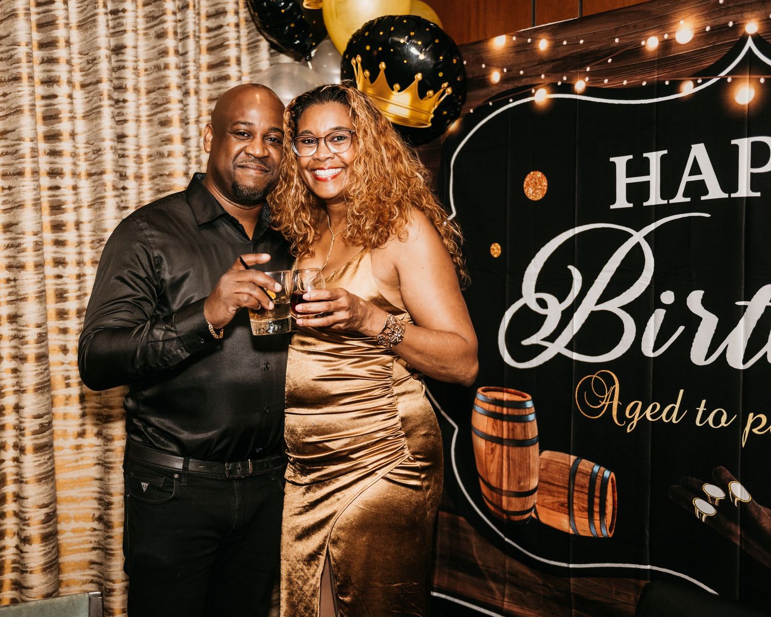 Couple posing together at a birthday party in front of a Happy Birthday backdrop, dressed in elegant black and gold attire.