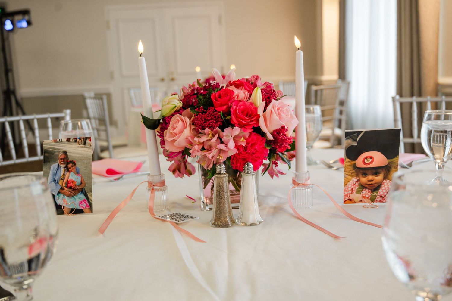 Elegant wedding reception table with pink and red floral centerpiece, tall white candles, and framed photos on white linen.