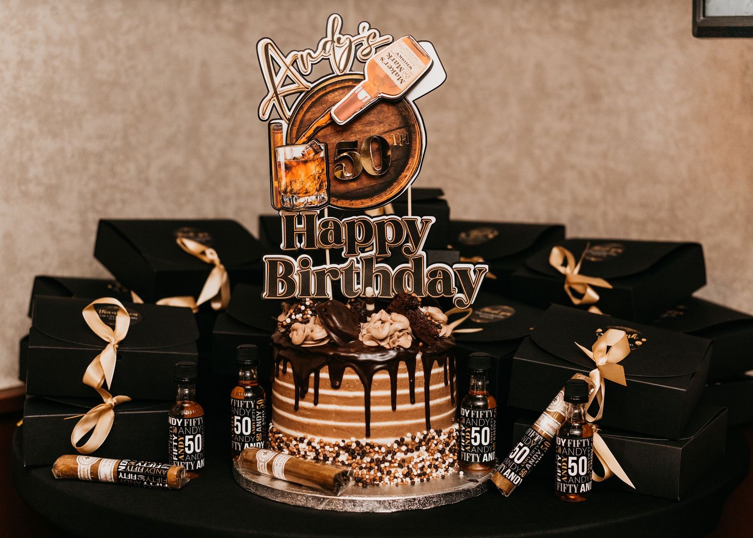 Elegant 50th birthday cake with chocolate drip, whiskey bottle decorations, and black gift boxes with gold bows.