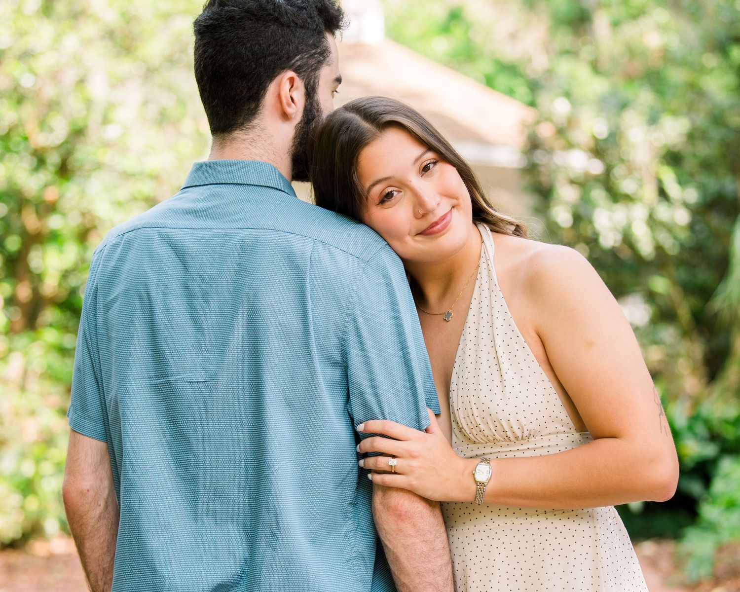 A woman in a floral dress leans on a man in a blue shirt outdoors, smiling warmly at the camera.