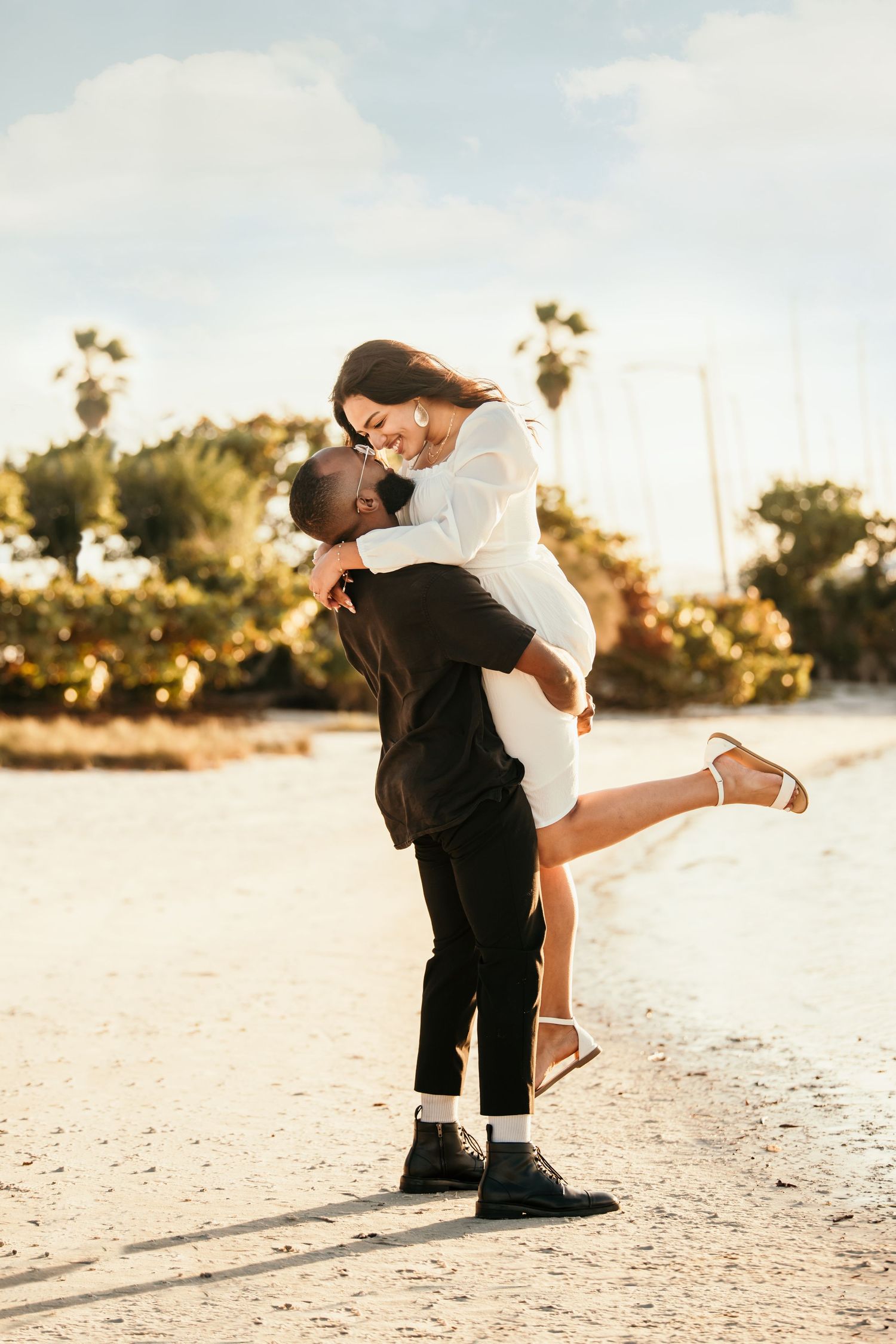 A couple shares a romantic embrace on a sunny beach, with the man lifting the woman in a white dress near palm trees.