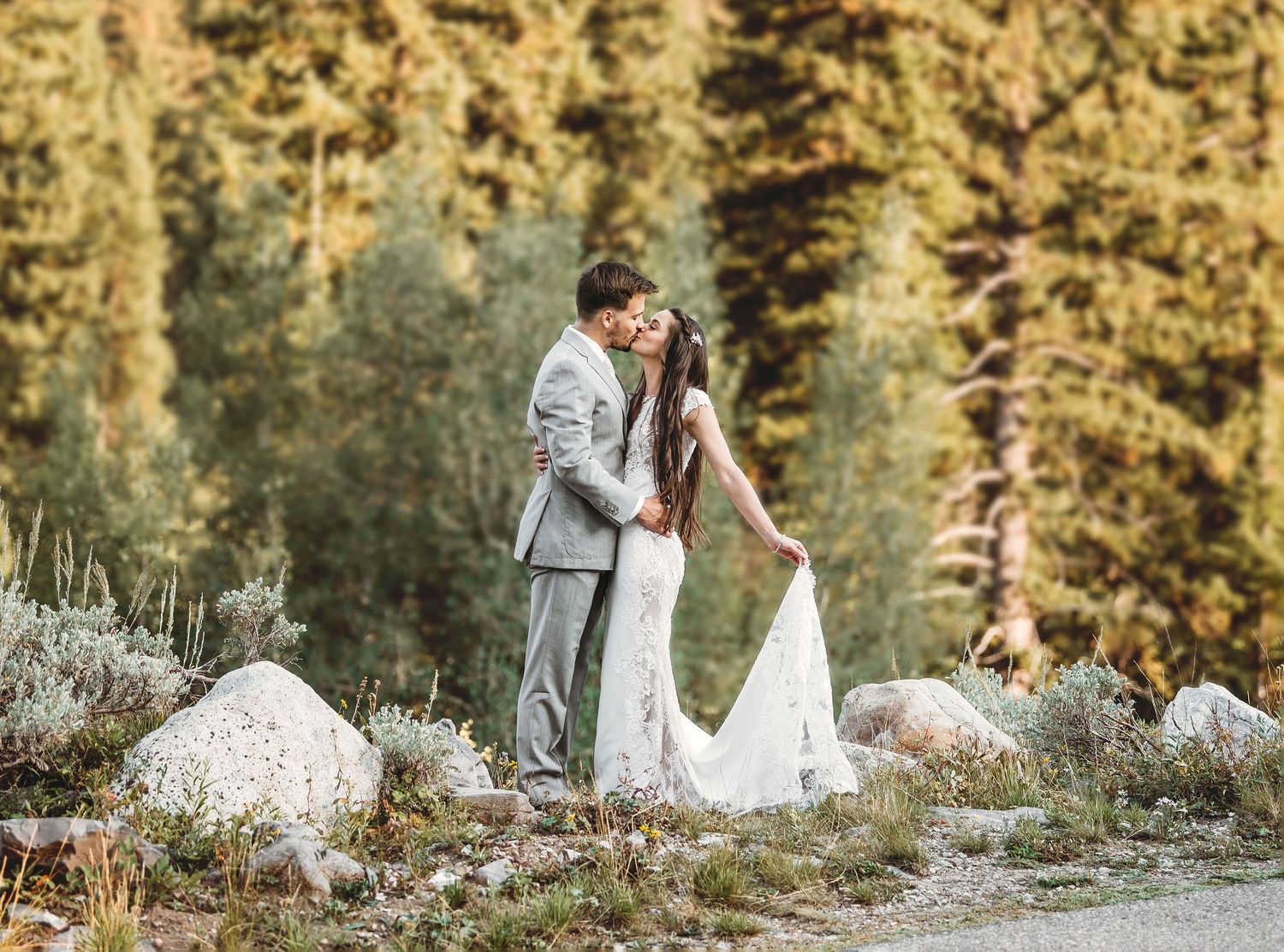 A couple shares a romantic kiss in wedding attire surrounded by lush green trees and rocky terrain outdoors.