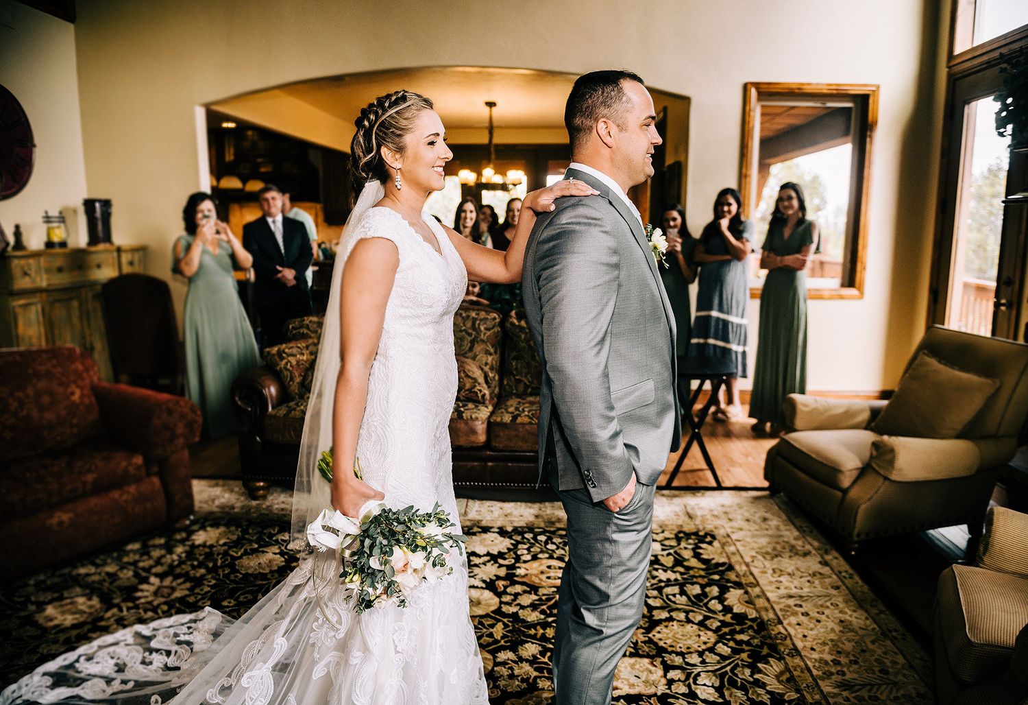 Bride in white gown holds bouquet during emotional first look moment with groom in gray suit at indoor wedding venue.