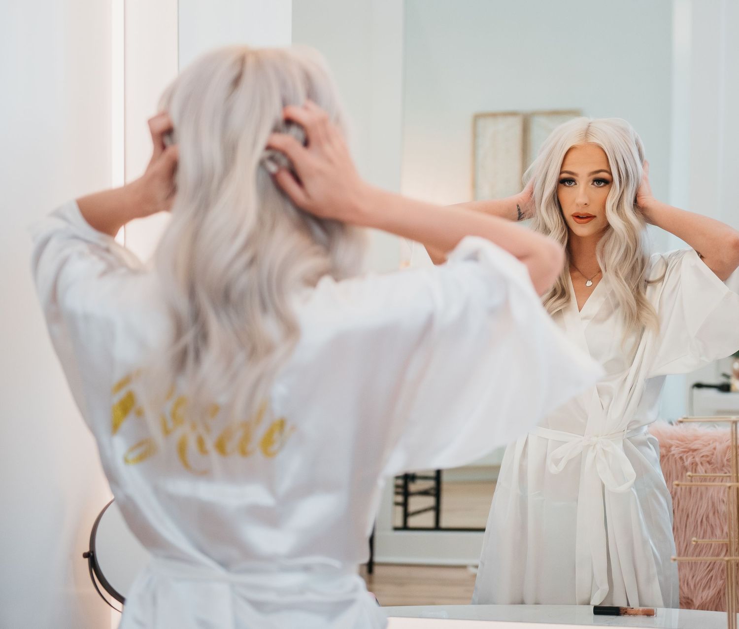 Bride in white robe with long blonde hair adjusting her hair while looking at her reflection in a mirror.
