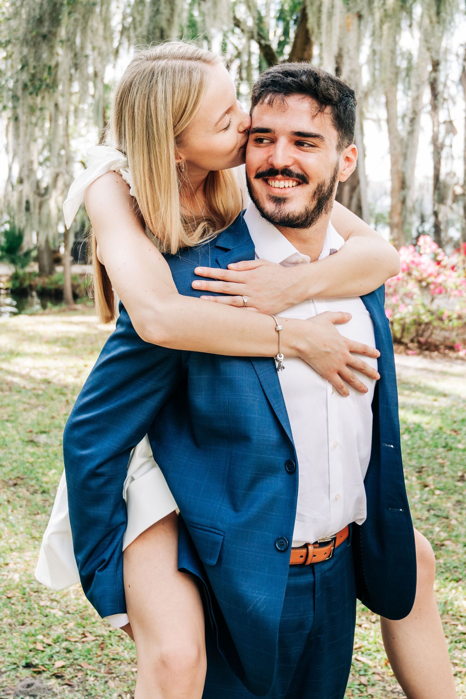 A smiling groom in a blue suit carries his blonde bride piggyback style outdoors as she kisses his cheek.