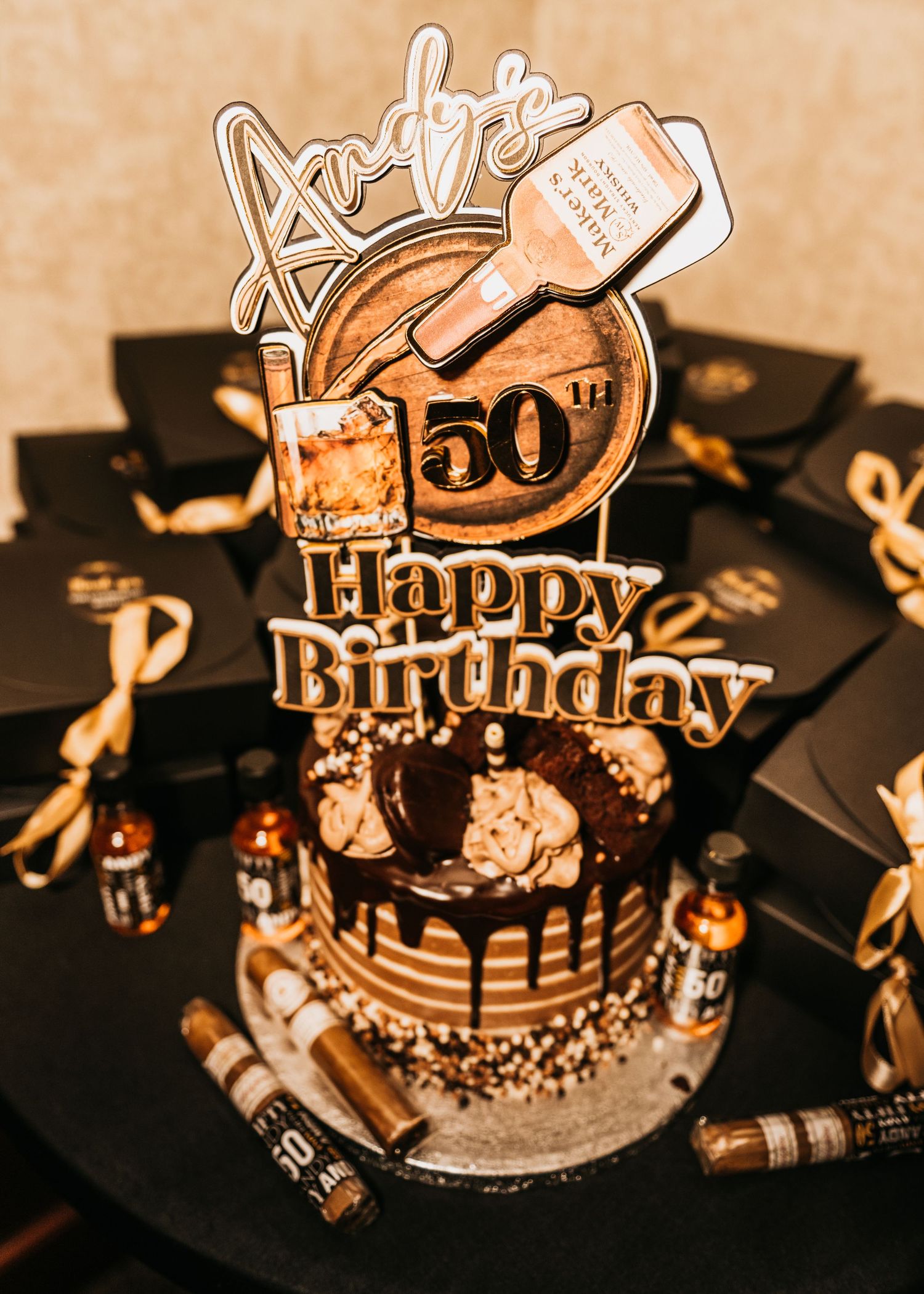 Andy's 50th Happy Birthday cake with gold drip frosting, cigar decorations, and whiskey-themed topper on a decorated table.