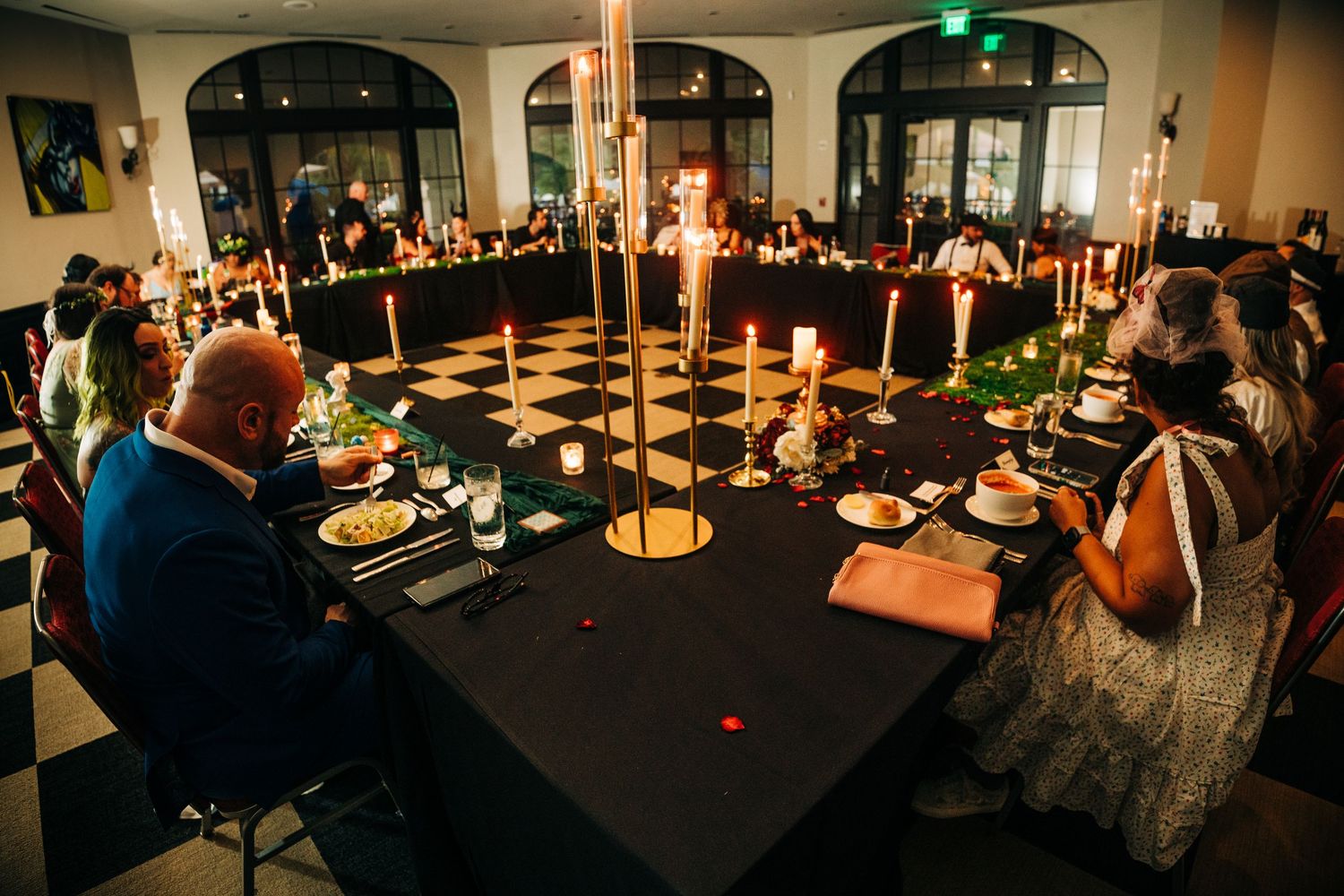 Elegant candlelit dinner party with guests seated at a large round table on a checkered floor in a dimly lit venue.