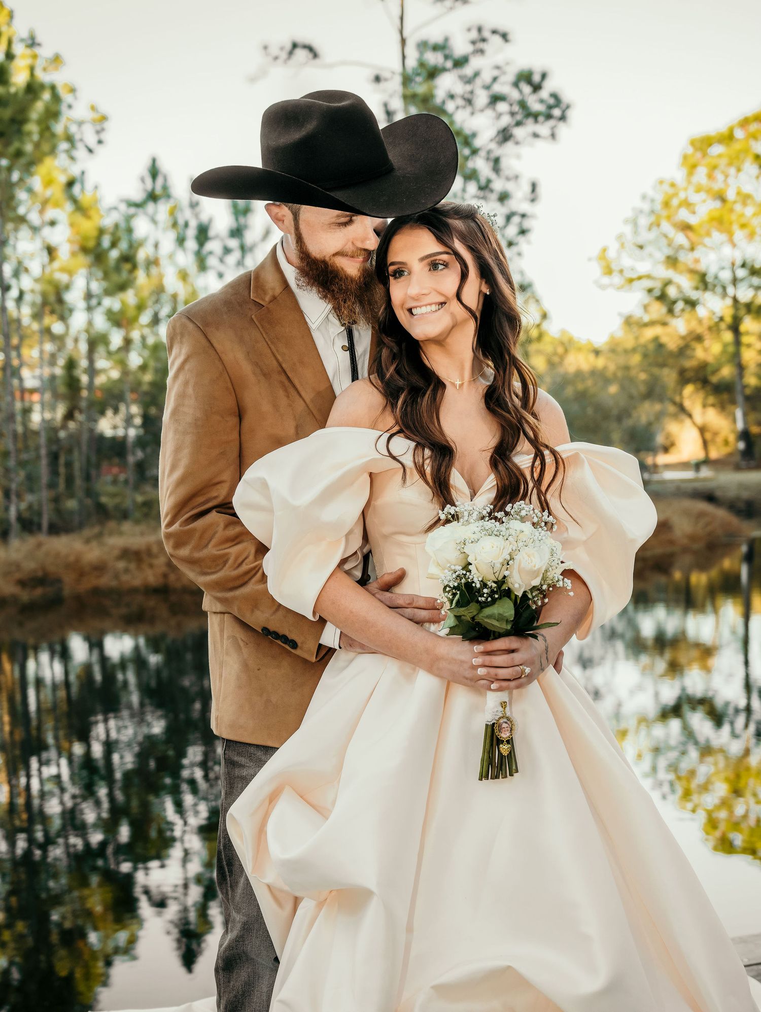 Bride in off-shoulder gown holds white bouquet beside groom in cowboy hat and tan suit near a pond.