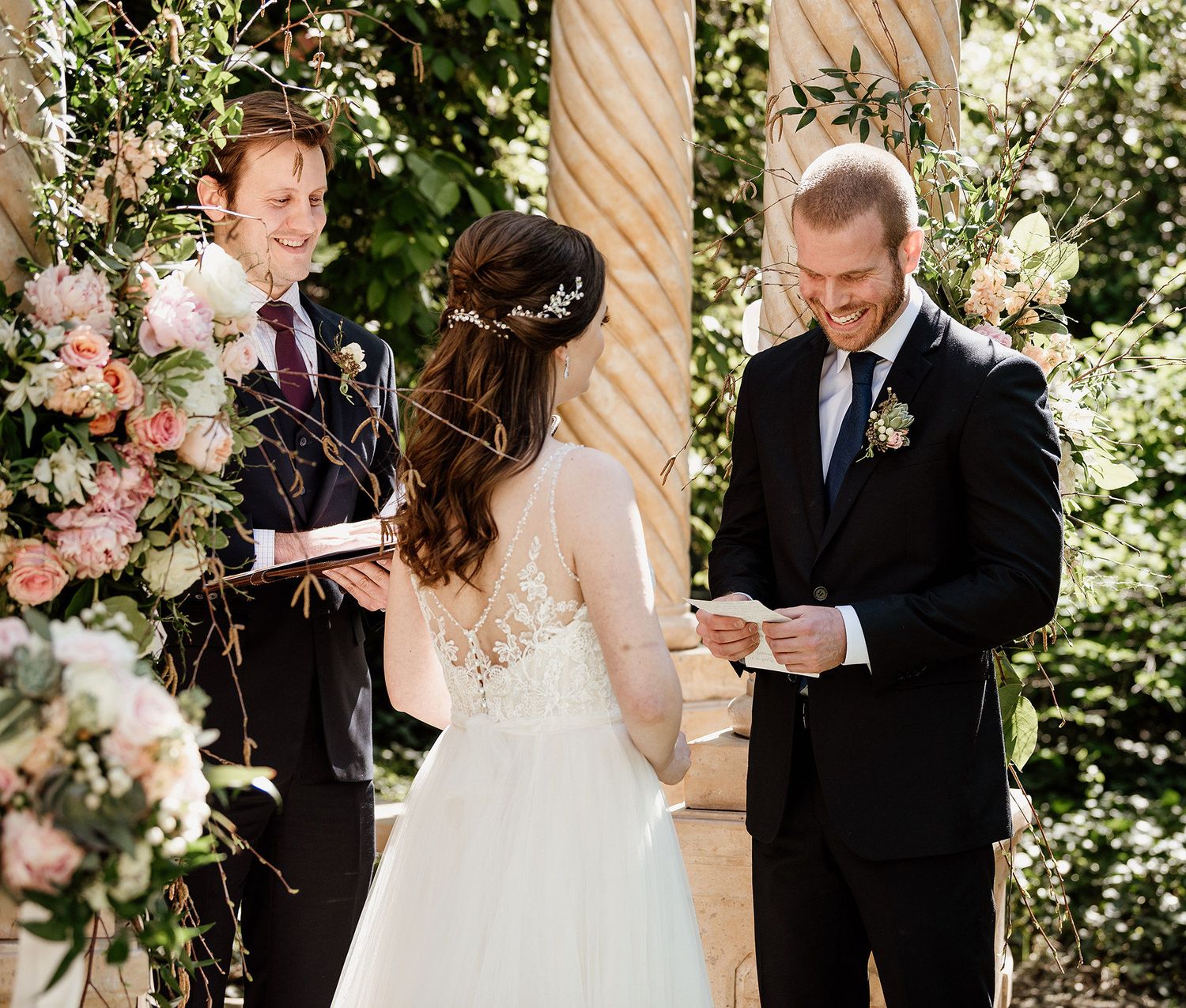 Groom reads wedding vows to bride in white lace dress at outdoor ceremony with floral arrangements.