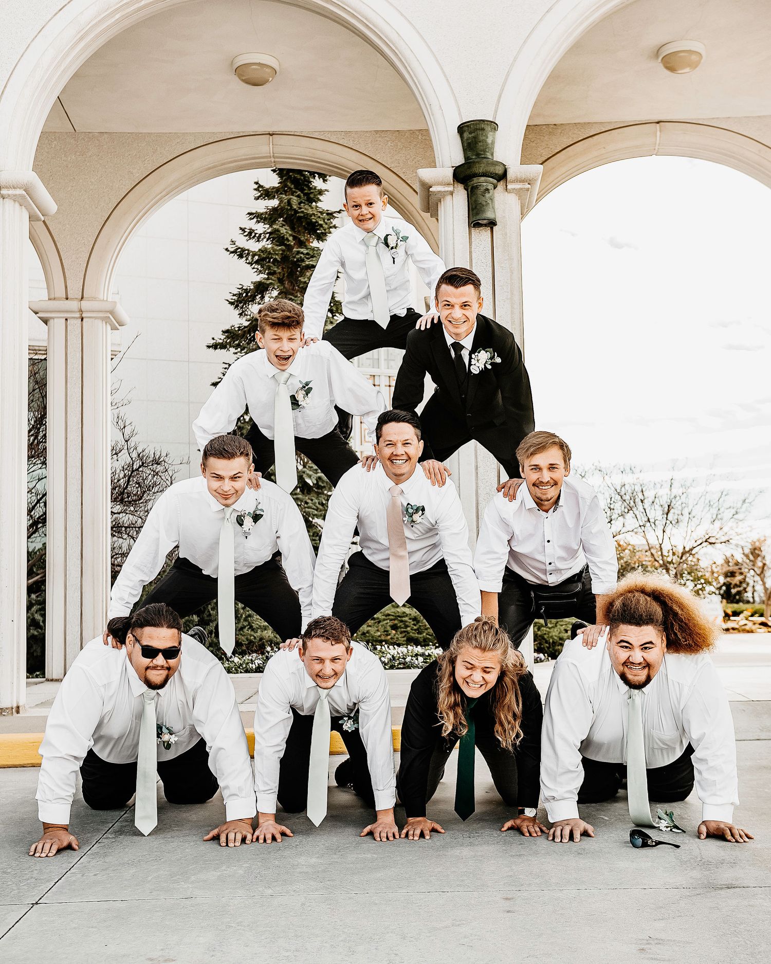Groomsmen forming a human pyramid in white shirts and black pants outside a wedding venue.