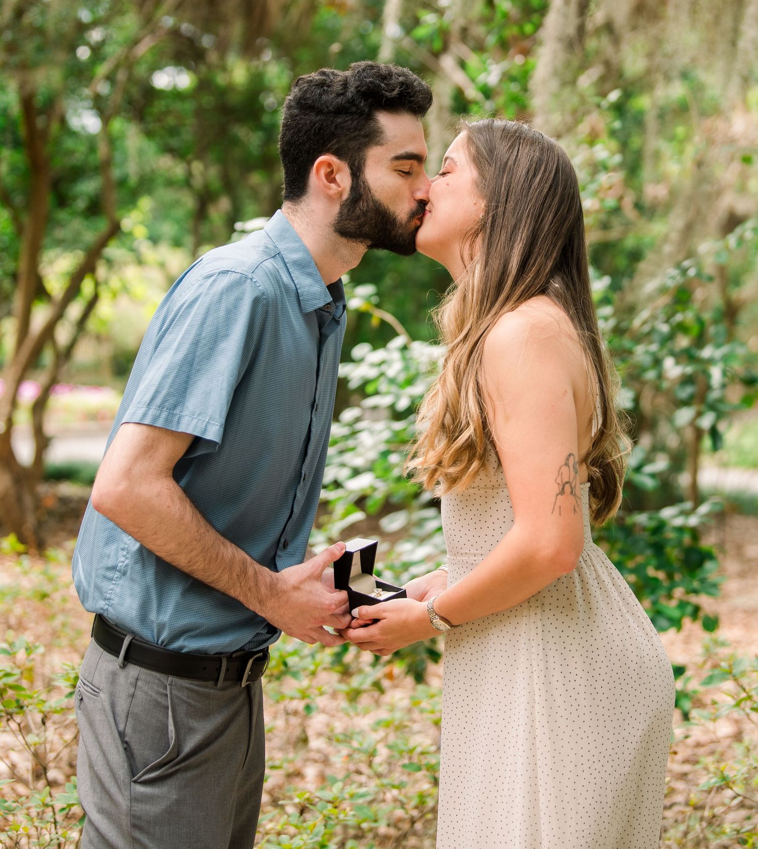 Couple sharing a kiss during an outdoor marriage proposal, man holding open ring box in a lush green park setting.