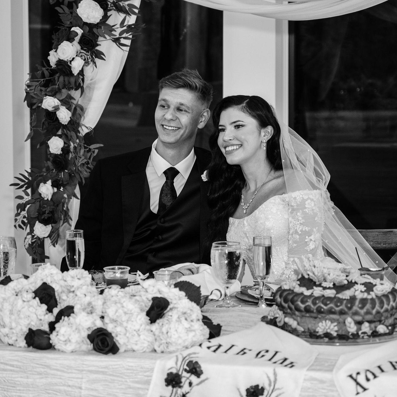 Black and white photo of a smiling bride and groom seated at their wedding reception table with floral arrangements.