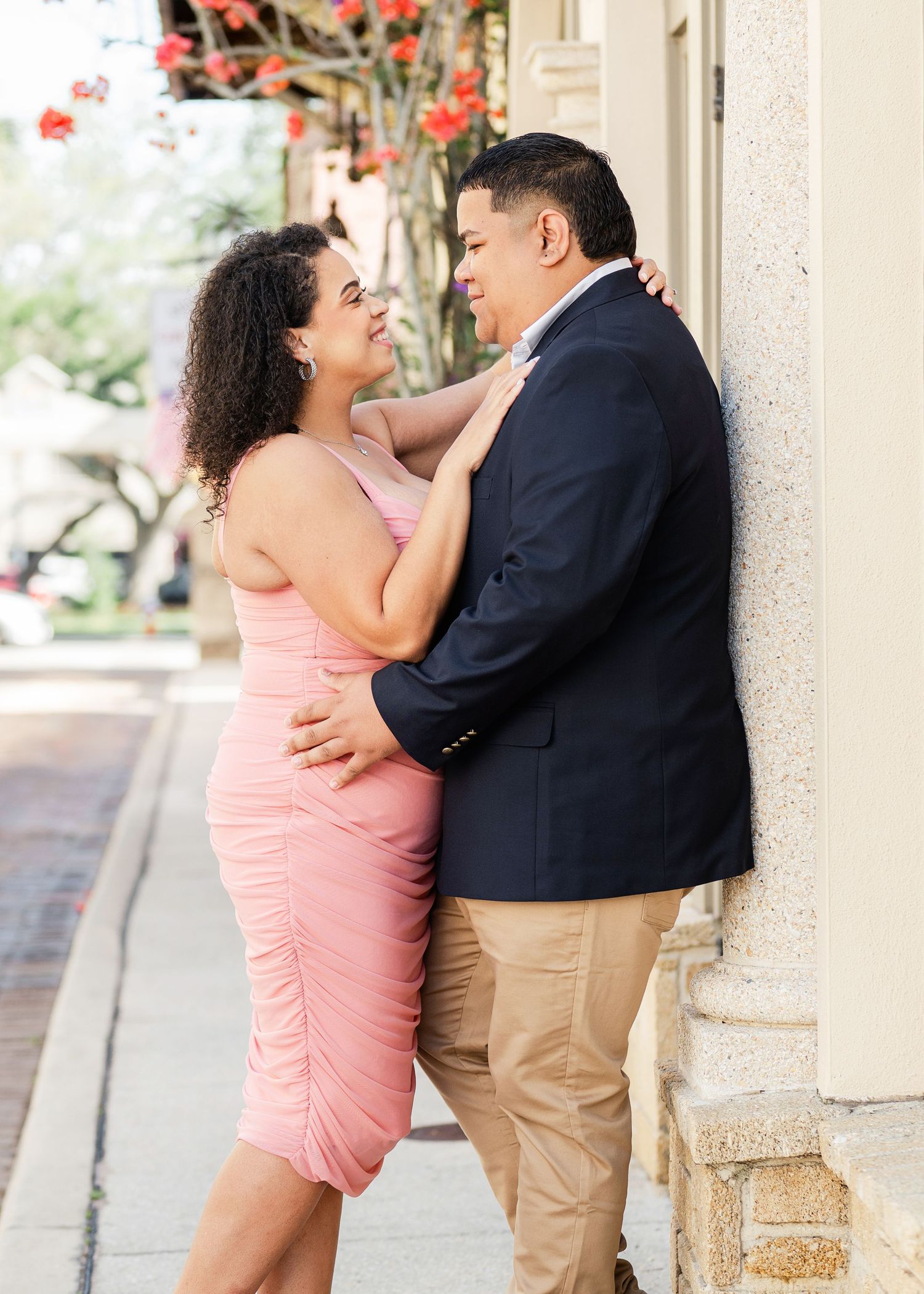 Couple in romantic embrace against a wall, woman in pink dress and man in navy blazer, sharing a tender moment outdoors.