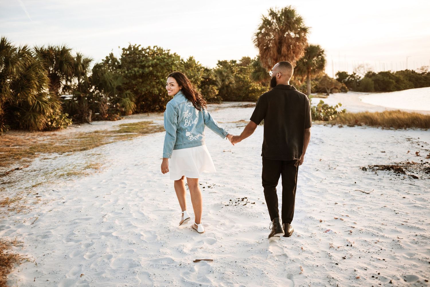 Couple holding hands walking on white sand beach at sunset with palm trees in background.