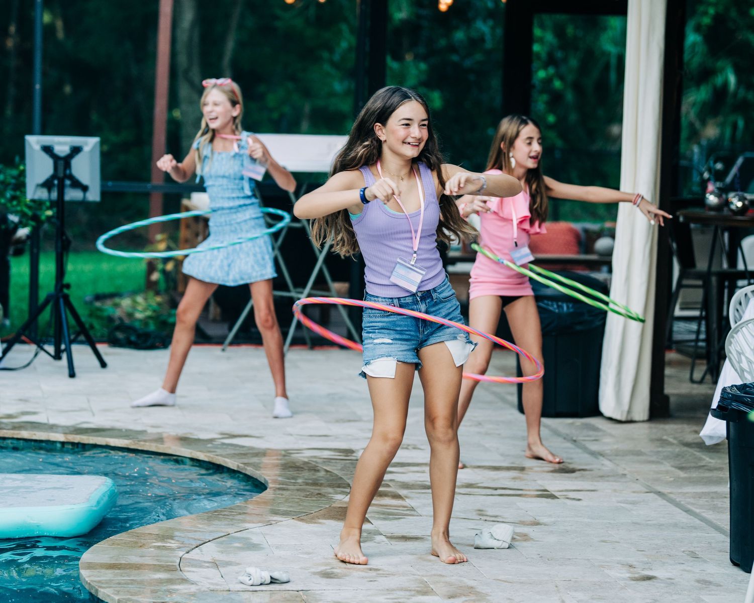Three girls hula hooping by a pool at an outdoor party on a patio.