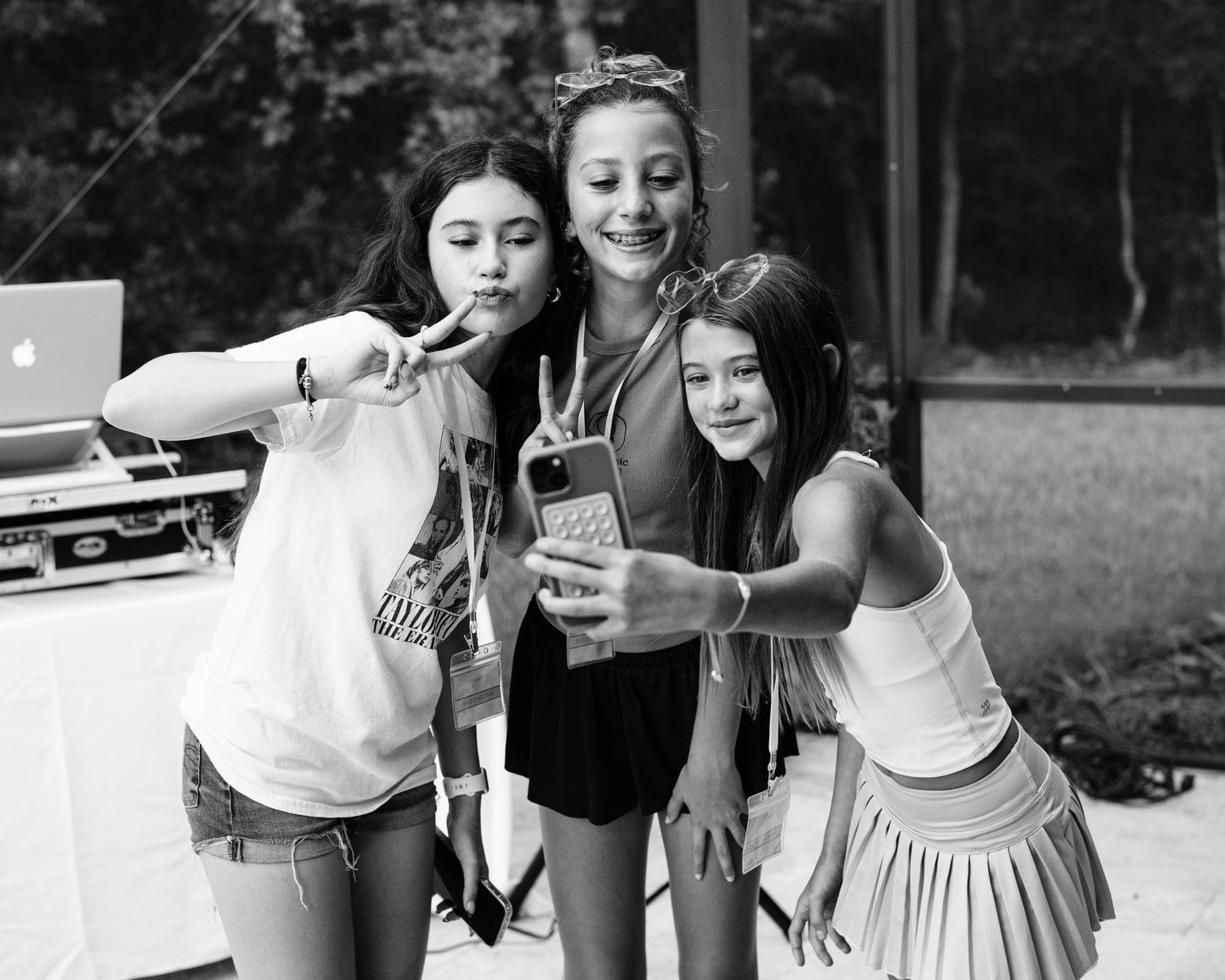 Three young girls taking a selfie together outdoors at a party, making peace signs in black and white photo.