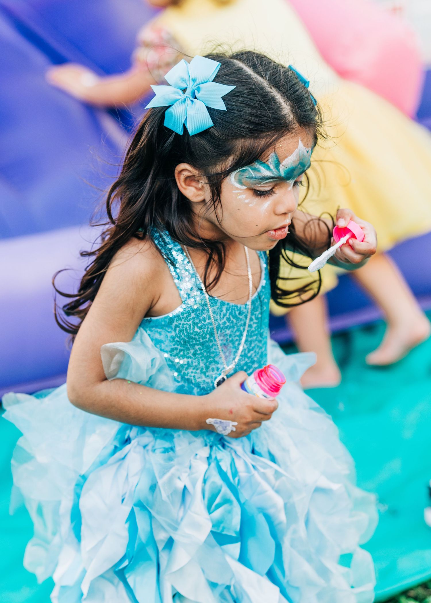 A young girl in a blue sequin dress with face paint and a bow blows bubbles at a festive outdoor party.