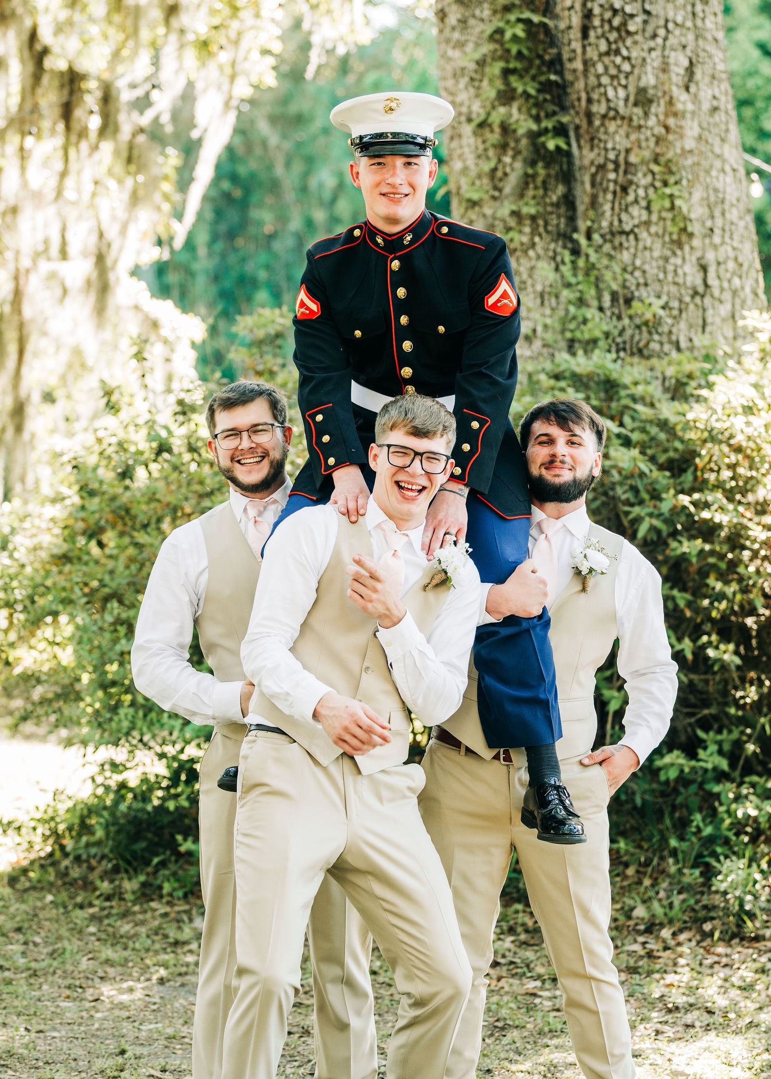 Groomsmen in tan suits pose playfully with a Marine in dress uniform at an outdoor wedding ceremony.