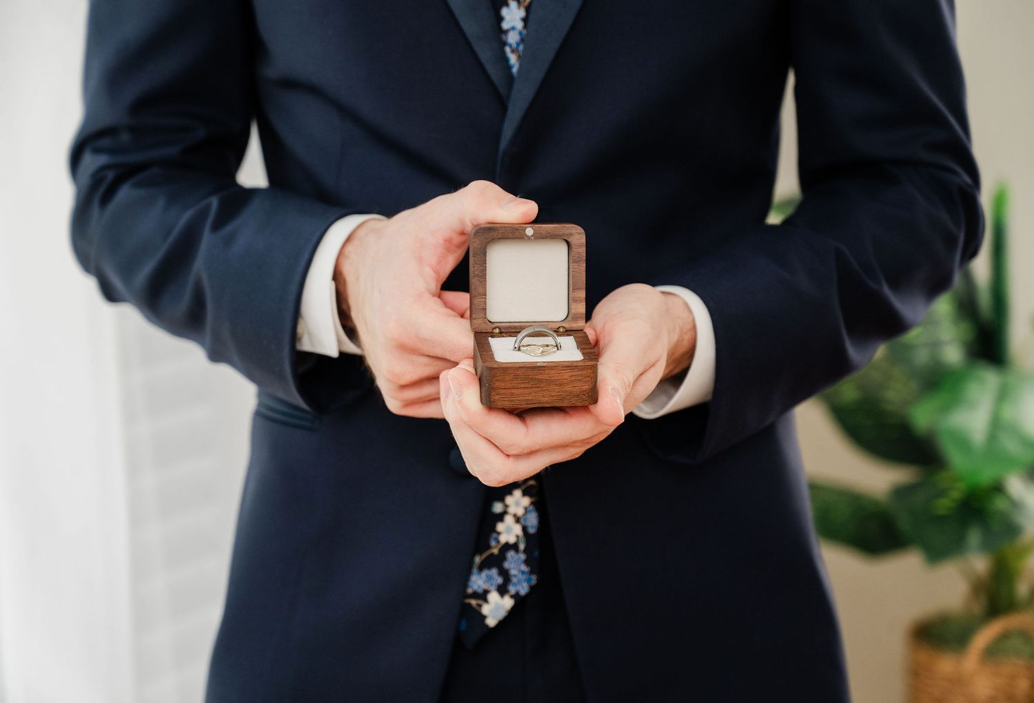 Groom in navy suit holding open wooden ring box with wedding rings at wedding ceremony.