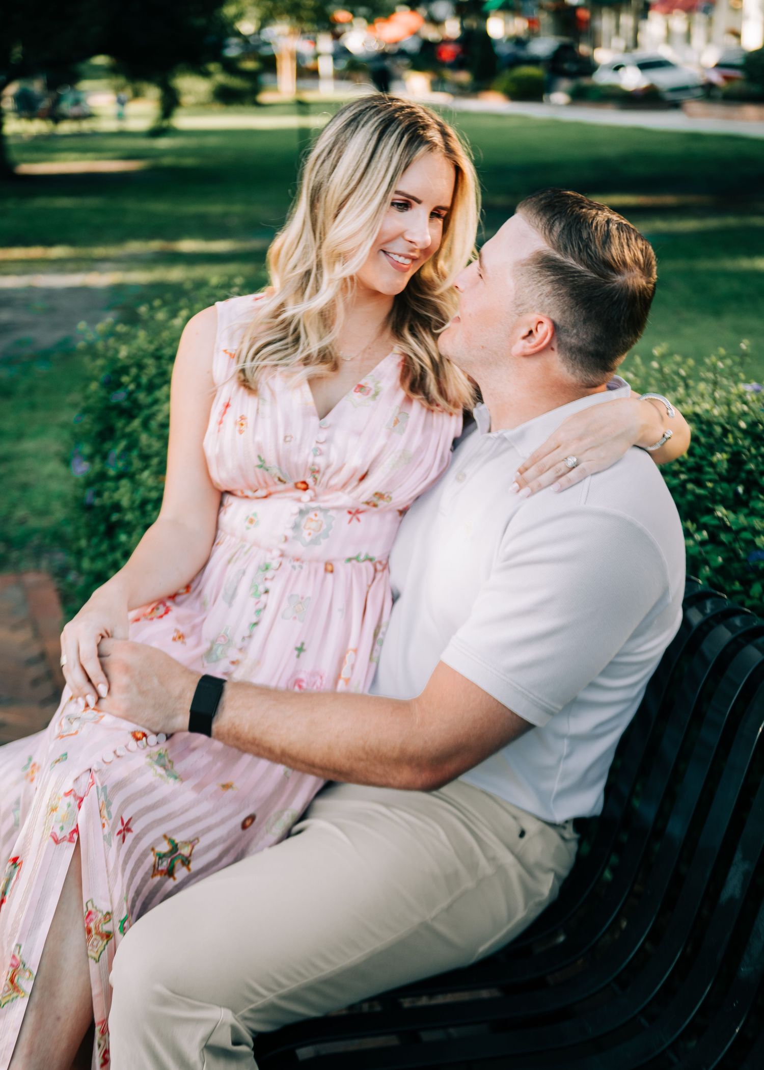 Couple sitting on a bench in a park, woman in floral pink dress with baby bump, man in white shirt embracing her.