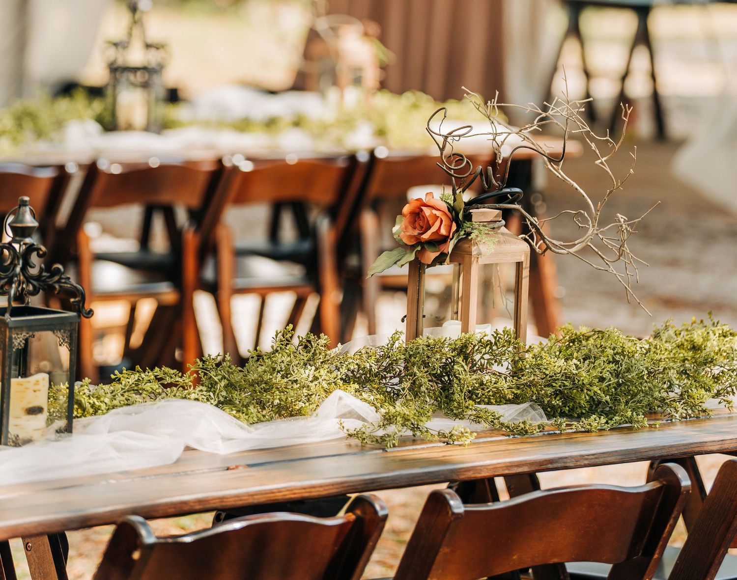 Elegant outdoor wedding reception table adorned with moss runner, orange roses, lanterns, and rustic wooden chairs.