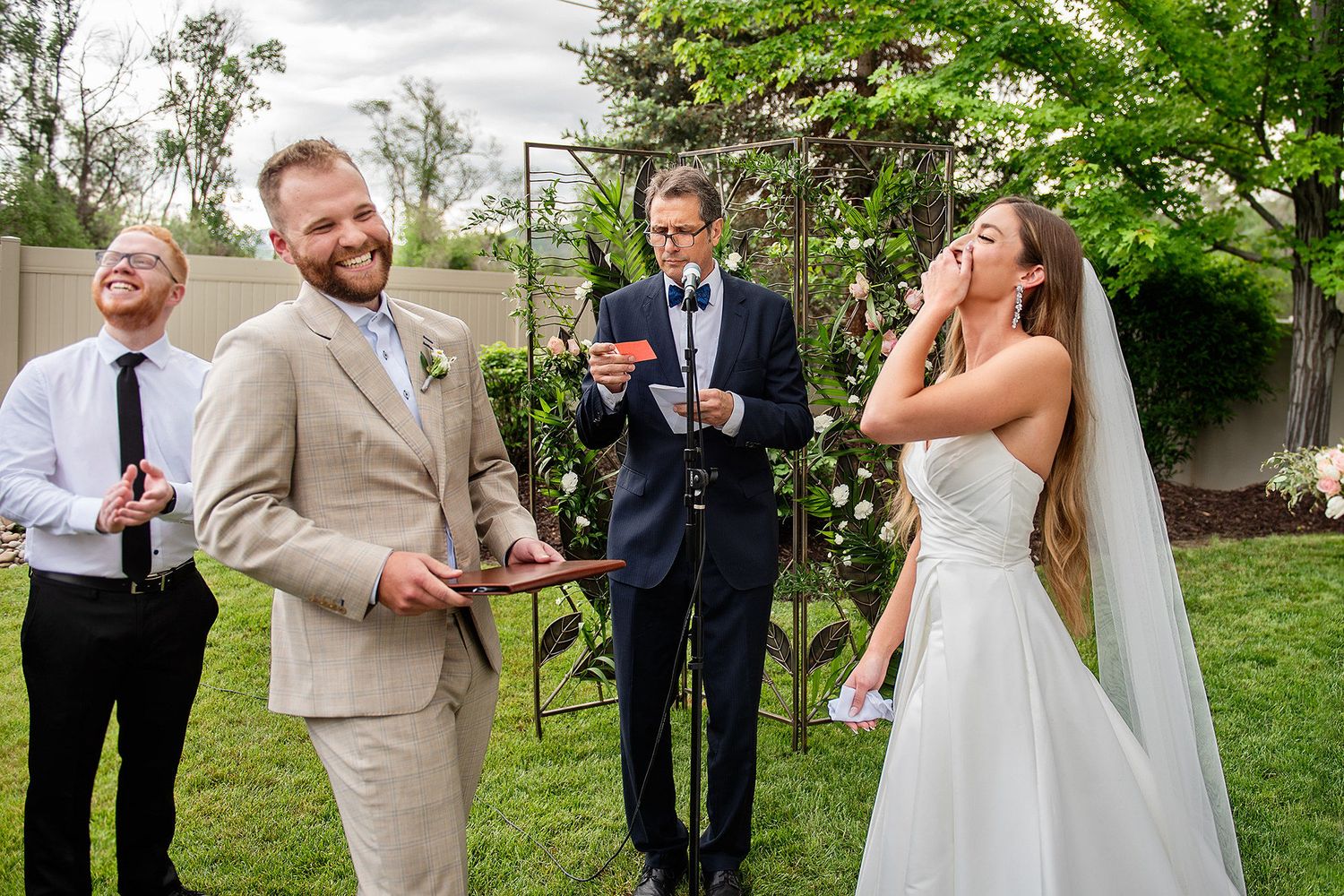 Outdoor wedding ceremony with laughing groom in tan suit, bride in white dress, and officiant reading vows in garden.