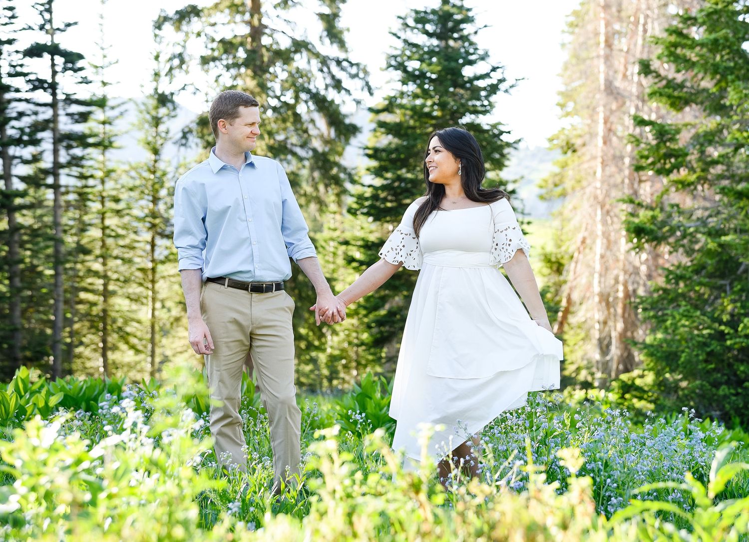Couple holding hands in a wildflower meadow surrounded by tall evergreen trees during an outdoor engagement photo session.